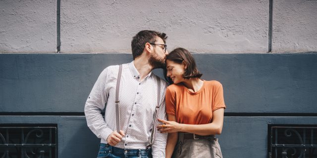 A white man wearing a white button-down shirt, blue jeans, and suspenders holds hands with and kisses a white woman with short brown hair wearing an orange top and a gray skirt.