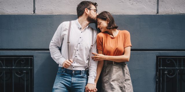 My Journey to Finding Love (and Marriage) as a Woman With a Learning Disability A white man wearing a white button-down shirt, blue jeans, and suspenders holds hands with and kisses a white woman with short brown hair wearing an orange top and a gray skirt.