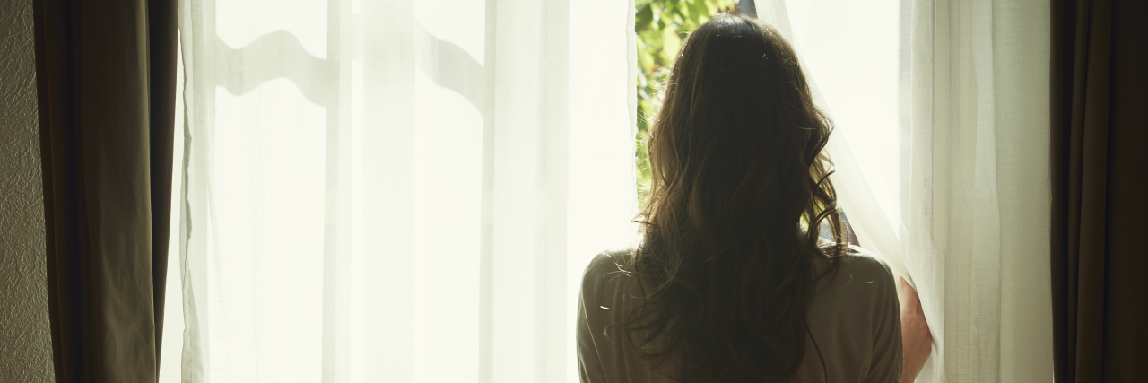 How Eating Disorder Treatment Can Both Help and Hurt You A woman with long brown hair wearing a soft pink top and jeans pulls back a curtain and looks out a window.