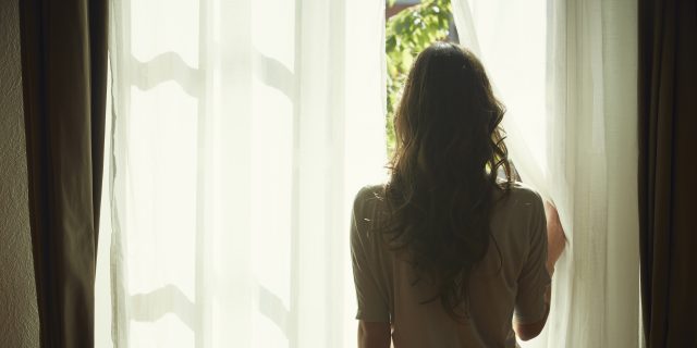 A woman with long brown hair wearing a soft pink top and jeans pulls back a curtain and looks out a window.