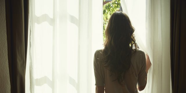 7 People With Eating Disorders Share What Eating Disorder Treatment Centers Do Well and How They Can Improve A woman with long brown hair wearing a soft pink top and jeans pulls back a curtain and looks out a window.