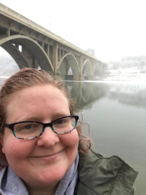 A white woman stands staring at the camera smiling with a snowy bridge behind her. She has glasses on and her hair pinned back with an olive green blazer.