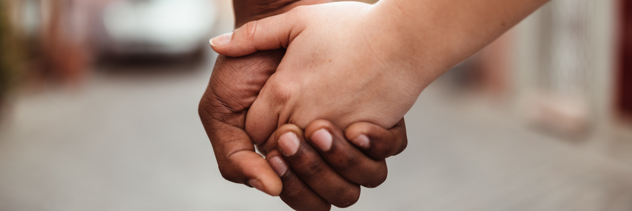 Close-up photo of a balck person and a white person holding hands in front of a street.