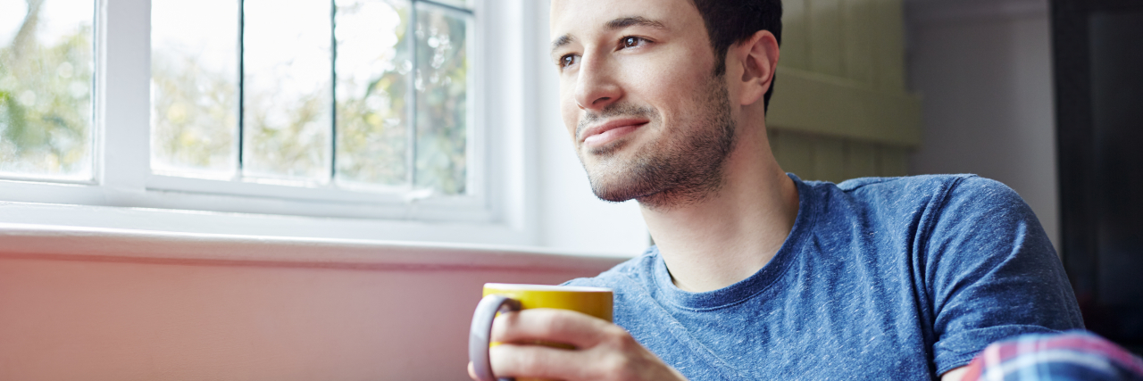 A white man with dark hair wearing a blue shirt and holding a mug sits on a red couch by a window.