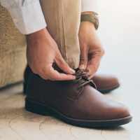 A man of color wearing a light blue button-down shirt, khaki pants, and brown shoes reaches down to tie his shoes.