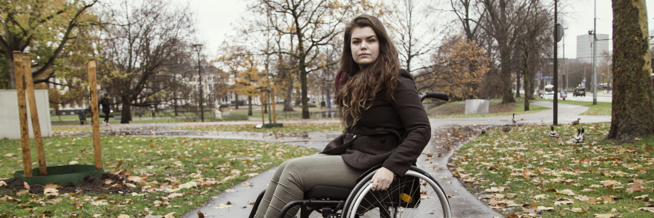 A woman with brown hair uses a wheelchair on a street during the fall. She is wearing a brown jacket.