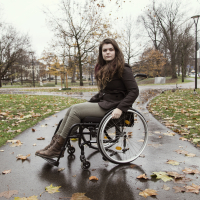 A woman with brown hair uses a wheelchair on a street during the fall. She is wearing a brown jacket.