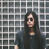 A man with long dark brown hair, a gray grunge-style T-shirt, a goth-style necklace, and a pair of sunglasses leans against a gray, latticed wall.
