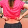 Back view of teenage girl with brown hair in a ponytail and wearing a pink T-shirt and black running shorts. She is holding her back in pain.