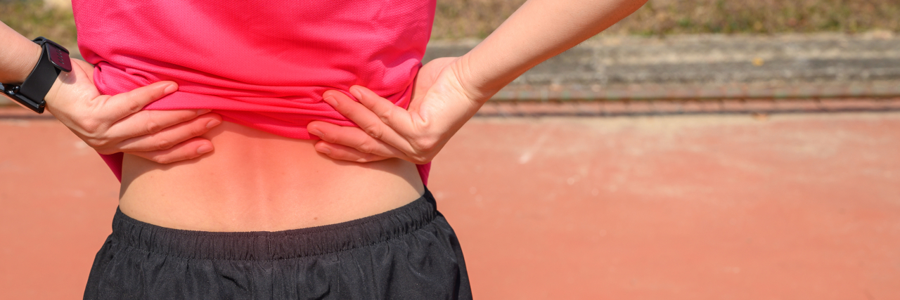 Surviving Life As a Teenager With Adolescent Idiopathic Scoliosis Back view of teenage girl with brown hair in a ponytail and wearing a pink T-shirt and black running shorts. She is holding her back in pain.