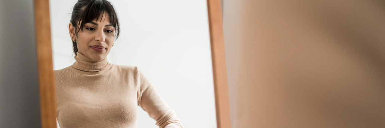 Affirmations for Entering 'Second Puberty' With an Eating Disorder a woman with dark brown hair and eyes wearing a soft pink shirt and jeans looks into the mirror while getting dressed.