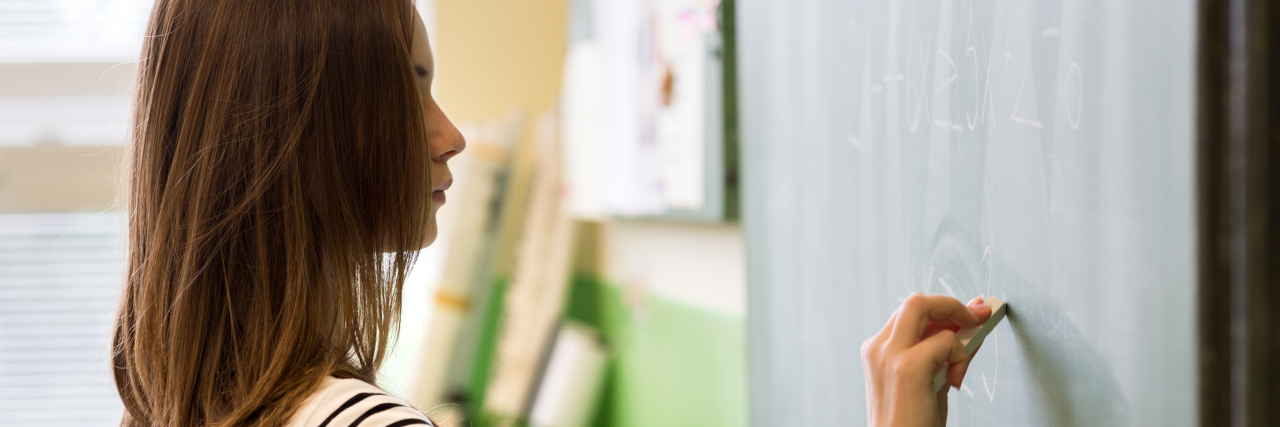 4 Coping Skills That Can Help Teachers With Their Mental Health A woman with long auburn hair wearing a black-and-white striped shirt writes on a blackboard while holding a folder.