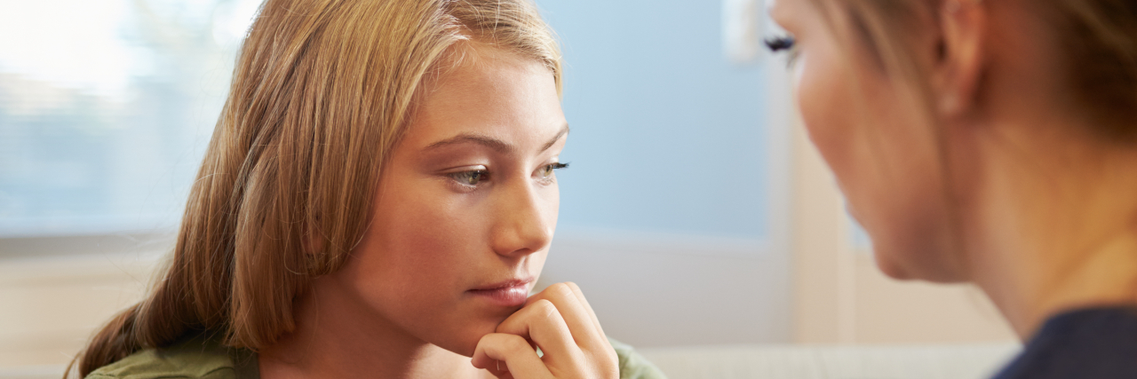 Having a Scoliosis Diagnosis and Surgery as a Young Teenager A blonde teen girl wearing a green V-neck T-shirt and a gray tank top underneath looks pensive while being comforted by a female nurse.