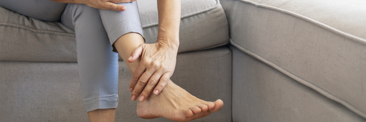 The Frustration of Living With the Lifelong Consequences of Medical Professionals' Mistakes A close-up photo of a woman wearing a pale blue top and pants holding her foot in pain.