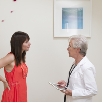 A woman with brown hair wearing a coral-colored dress stands across from an elderly female doctor in a doctor's office with her hands on her hips.