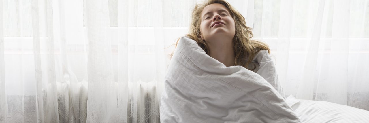 A Message for Your Hard Days With Bipolar Disorder A woman with strawberry-blonde hair sits on a bed while hugging herself with a comforter. She has her eyes closed and sits in front of a set of curtains.