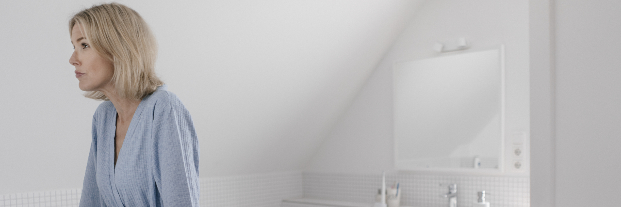 Getting Stuck and Being Resourceful With Cerebral Palsy A white woman with short blonde hair wearing a blue bathrobe leans against a counter and looks ahead in a white bathroom.