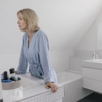 A white woman with short blonde hair wearing a blue bathrobe leans against a counter and looks ahead in a white bathroom.
