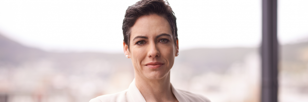 Accepting Myself With Schizophrenia and Letting Go of Shame A woman with short brown hair and brown eyes wearing a white shirt, necklace, and white blazer, stands confidently against a balcony.