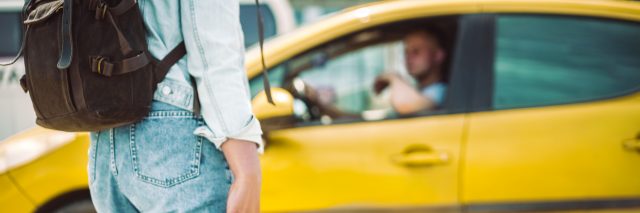 A person wearing a denim shirt and jeans holds onto the handle of a suitcase while looking out at a yellow car driven by a man.