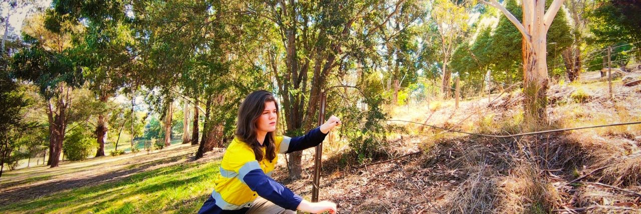 Working a Physical Job and Discrimination With Fibromyalgia The author, a white woman with curly brown hair wearing a yellow vest, blue undershirt, and jeans assesses the condition of a fence.