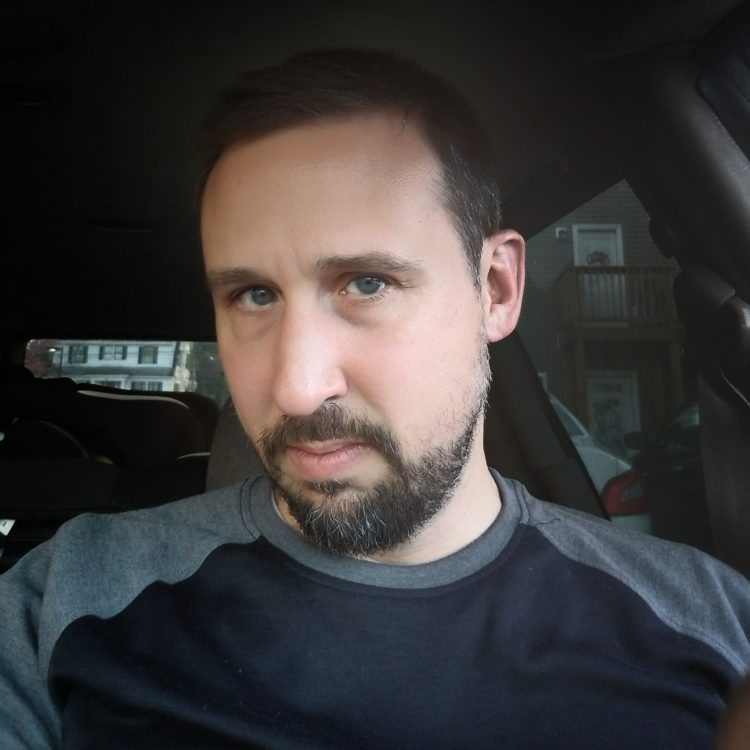 A white man with brown hair and blue eyes sits in a car and looks at the camera.