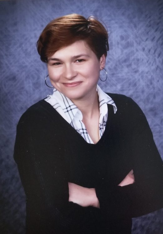 A white woman with short auburn hair crosses her arms while wearing a black sweater, a white plaid collard shirt, and small hoop earrings. She is standing in front of a blue background.