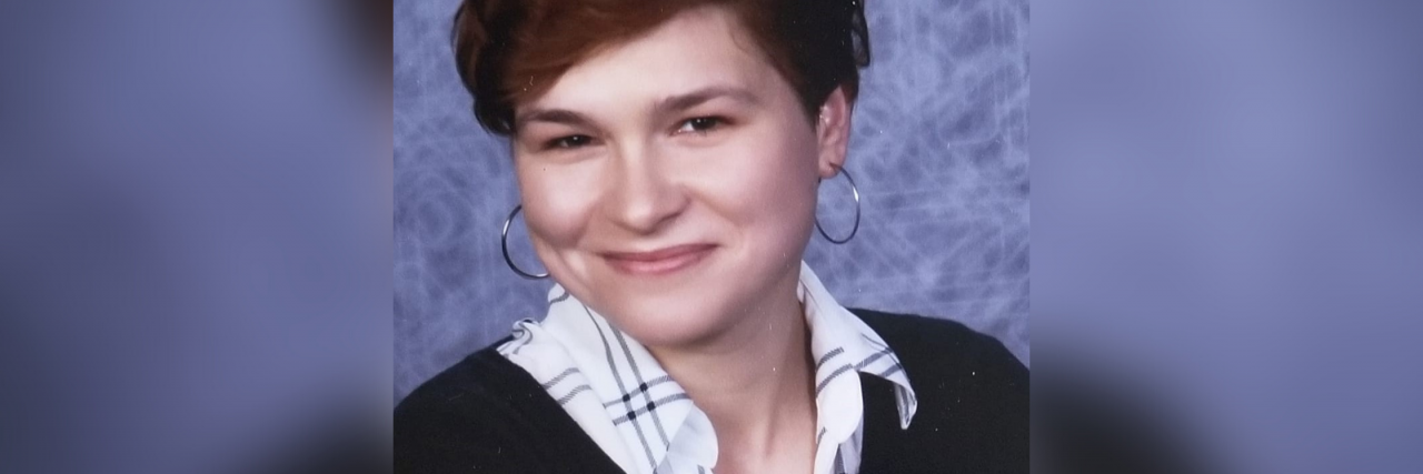 A white woman with short auburn hair crosses her arms while wearing a black sweater, a white plaid collard shirt, and small hoop earrings. She is standing in front of a blue background.