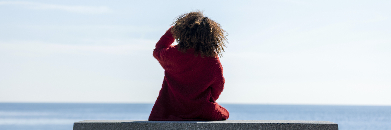 Rear view of a young black woman sitting on a bench while looking away to the horizon over the sea