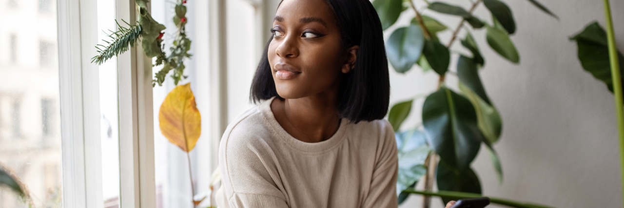 It's Possible to Live Alone If You Have Trauma or Anxiety Woman sitting on the windowsill with mobile phone and looking away. Woman relaxing at home looking outside the window.