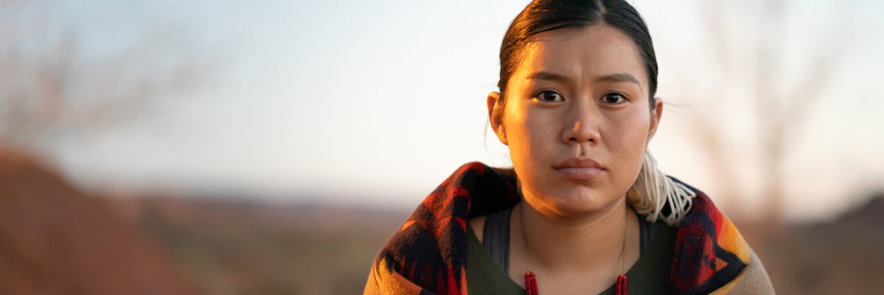 An indigenous woman looking straight ahead. She has a colorful blanket wrapped around her, and is standing outside at sunset