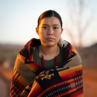 An indigenous woman looking straight ahead. She has a colorful blanket wrapped around her, and is standing outside at sunset