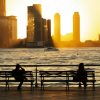 Silhouette of two people sitting on park benches in front of river and skyline at sunset
