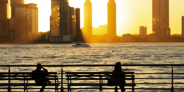 Silhouette of two people sitting on park benches in front of river and skyline at sunset