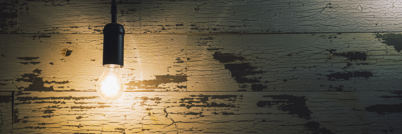 Illuminating the Dark Side of Life With Chronic Illness Vintage light bulb hanging against weathered wooden wall in the darkness.