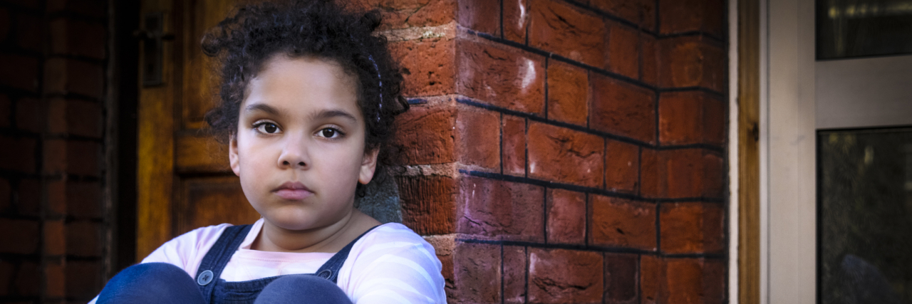 Little girl with knees tucked to her chest sitting against a brick wall, sad