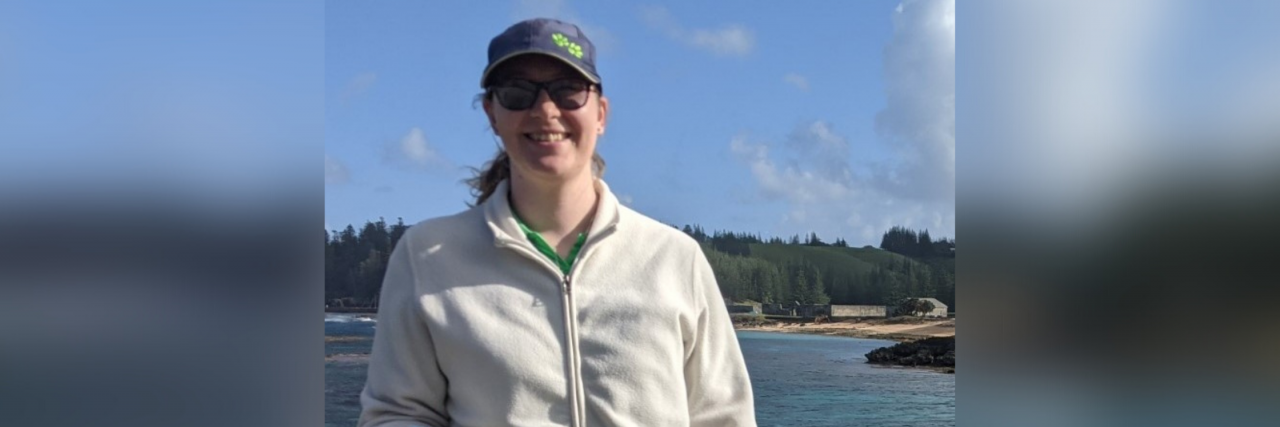 What Helps a Woman in Her Thirties With Rheumatoid Arthritis A woman wearing a white jacket, black pants, a navy blue baseball cap, and sunglasses who's holding a walking stick stands in front of a lake, smiling.