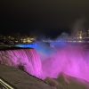 The picture is taken at night with a dark sky. There is water rushing over the falls and mist which floated up from the rocks below. There is a light shining on the water that makes it look like the water is pink, green and bleu in honor of Rare Disease Day. There is a shyline behind the lit up falls with both low and tall buildings of different shapes. The buildings have lit white/yellow rectangle windows. There is a tall building with the word CASINO lit on top of the building in red.