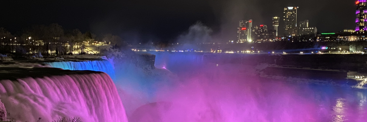 The picture is taken at night with a dark sky. There is water rushing over the falls and mist which floated up from the rocks below. There is a light shining on the water that makes it look like the water is pink, green and bleu in honor of Rare Disease Day. There is a shyline behind the lit up falls with both low and tall buildings of different shapes. The buildings have lit white/yellow rectangle windows. There is a tall building with the word CASINO lit on top of the building in red.