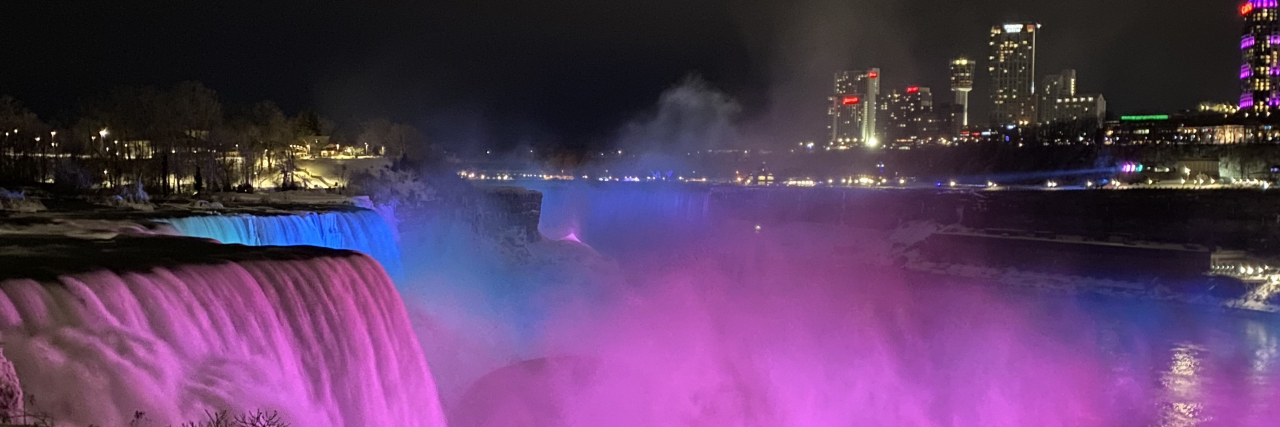 Why the Illumination of the Rare Disease Community Is So Important The picture is taken at night with a dark sky. There is water rushing over the falls and mist which floated up from the rocks below. There is a light shining on the water that makes it look like the water is pink, green and bleu in honor of Rare Disease Day. There is a shyline behind the lit up falls with both low and tall buildings of different shapes. The buildings have lit white/yellow rectangle windows. There is a tall building with the word CASINO lit on top of the building in red.