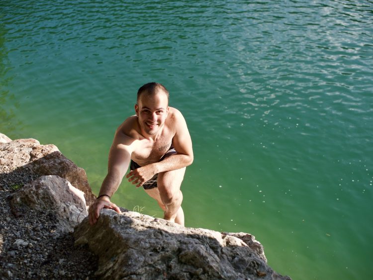 Author: Young man in swimming trunks standing on a rock above the water, smiling brightly