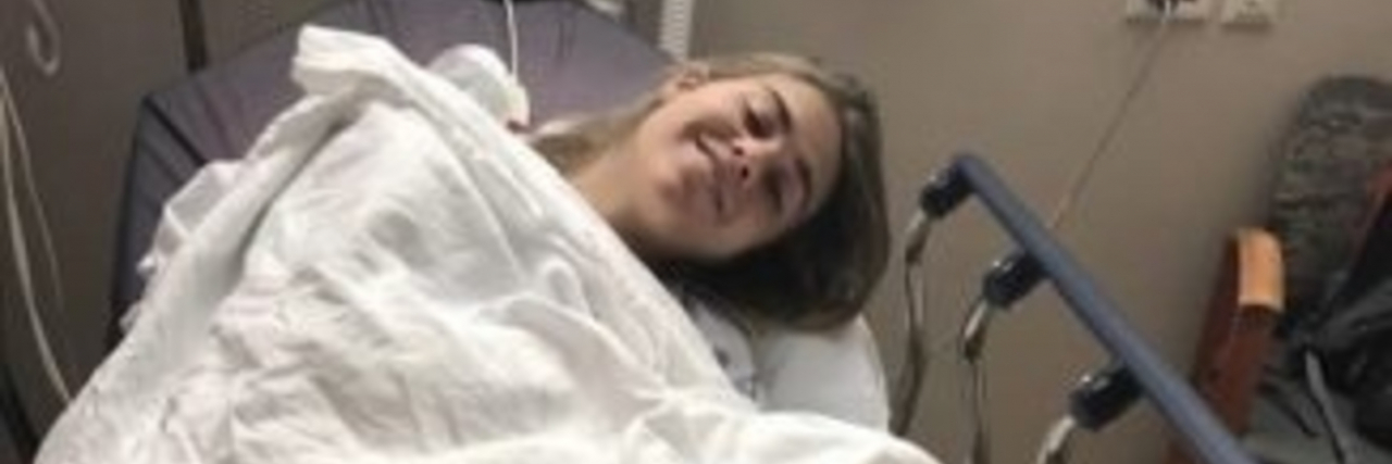 How My Ehlers-Danlos Syndrome Contributed to My Eating Disorder The author, a white woman with short brown hair, lies in a hospital bed covered by a sheet alongside her brown dog.