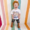 The author's son with Down syndrome stands against a colorful, striped background while wearing a white graphic t-shirt, long pants, and brown boots and smiling brightly.