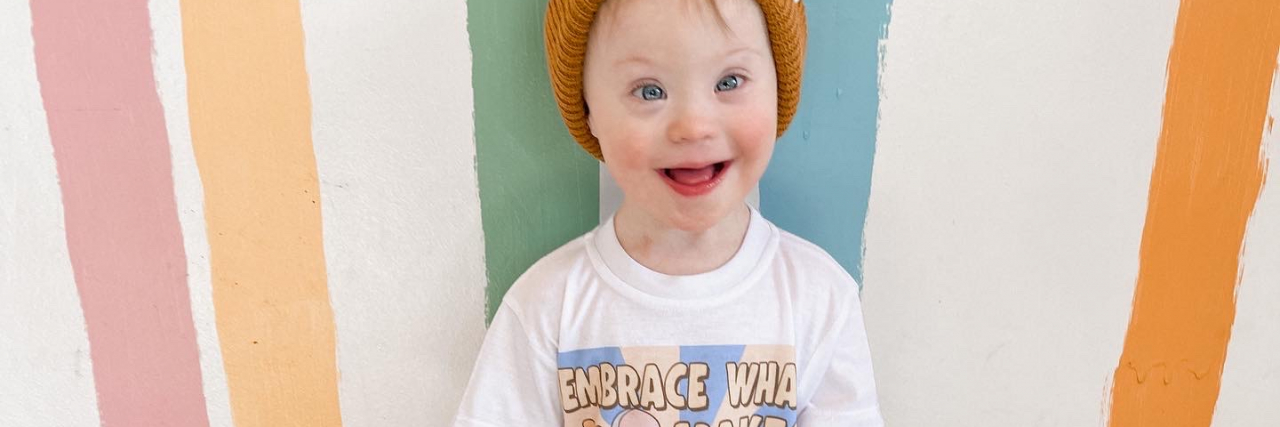 Being Part of the 'Lucky Few' While Raising a Child With Down Syndrome The author's son with Down syndrome stands against a colorful, striped background while wearing a white graphic t-shirt, long pants, and brown boots and smiling brightly.