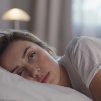 A woman with blonde hair sleepily looks into teh camera as she lies awake in bed.