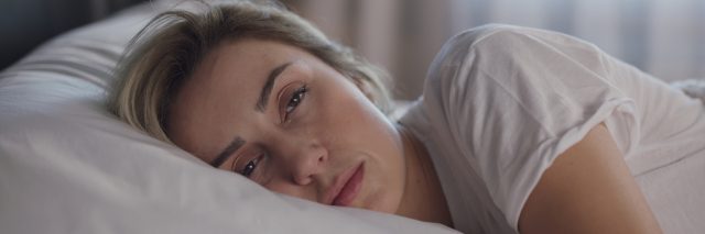 A woman with blonde hair sleepily looks into teh camera as she lies awake in bed.