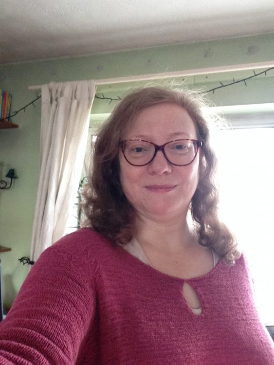 A white woman with curly red hair, blue eyes, and glasses, smiles softly at the camera while wearing a red top.