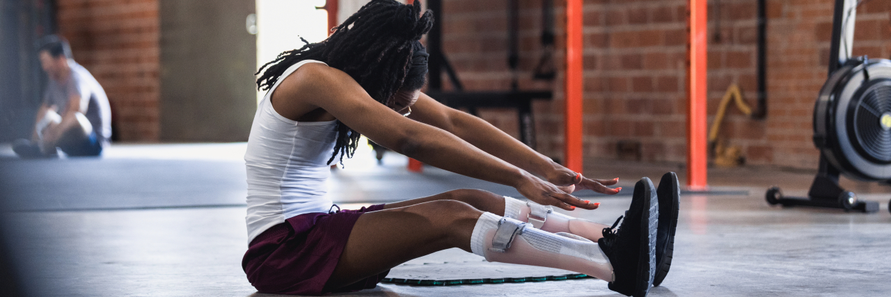 A black woman in a warehouse gym wearing a white tank top and purple athletic shorts stretches her legs and reaches towards her legs with her arms. She wears light pink leg braces.