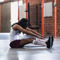 A black woman in a warehouse gym wearing a white tank top and purple athletic shorts stretches her legs and reaches towards her legs with her arms. She wears light pink leg braces.