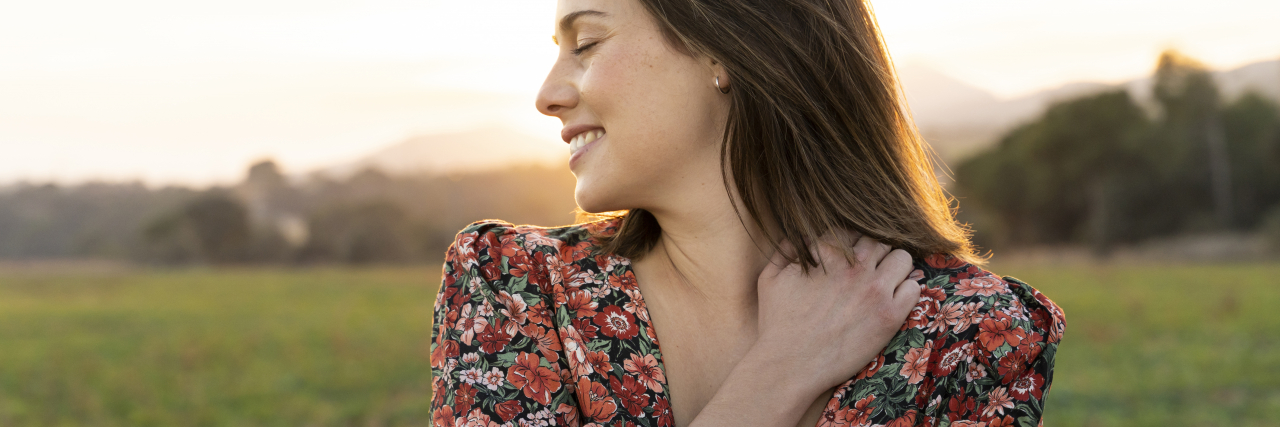 A white woman with brown hair wearing a floral blouse hugs herself outside.